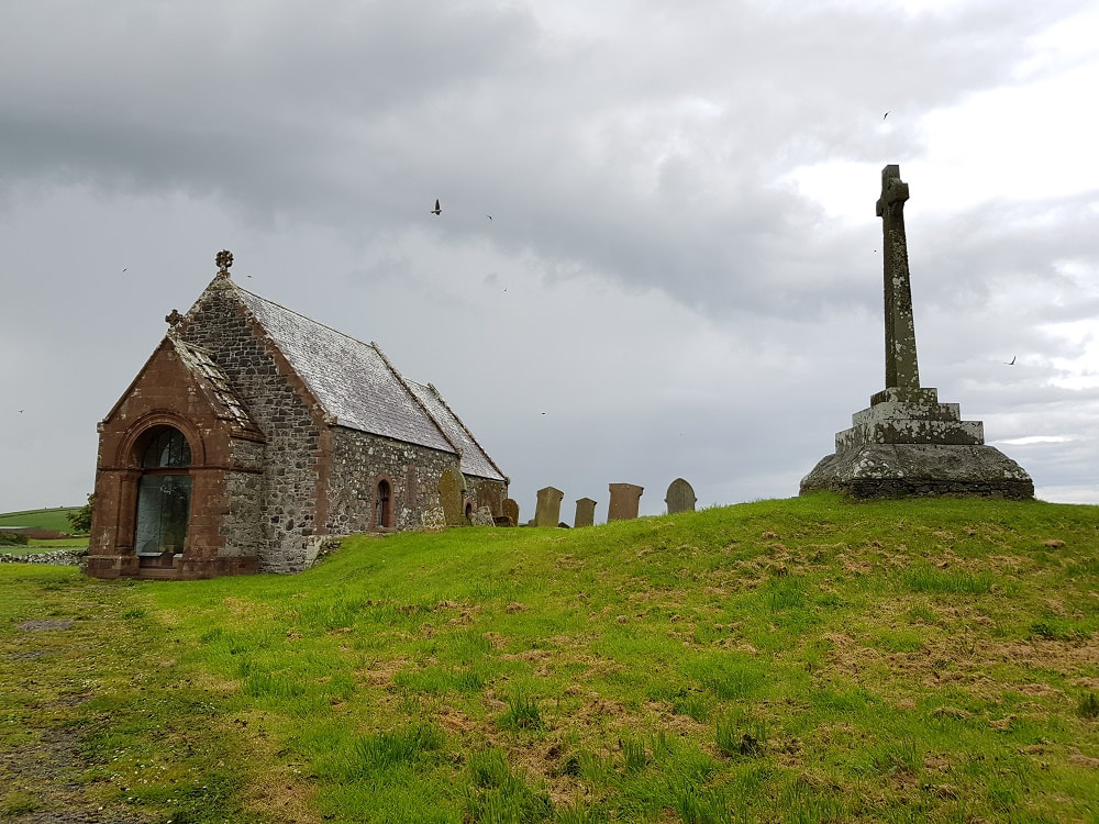PicturA brown stone church next to a tall grey stone cross, sitting on top of a green hill