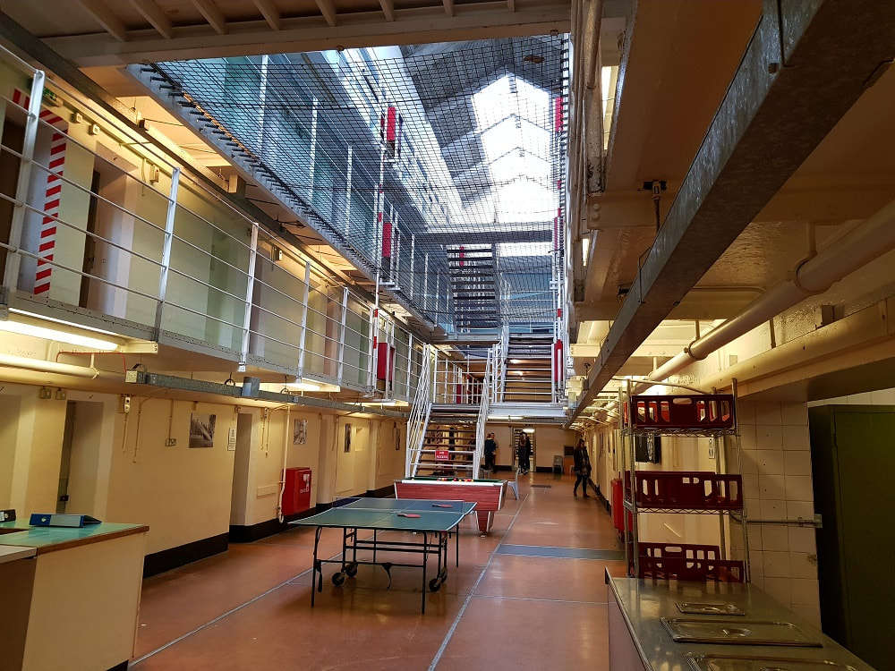 The inside of a prison building with three storeys of cells.  A table tennis set sits on the ground floor and several people are walking around