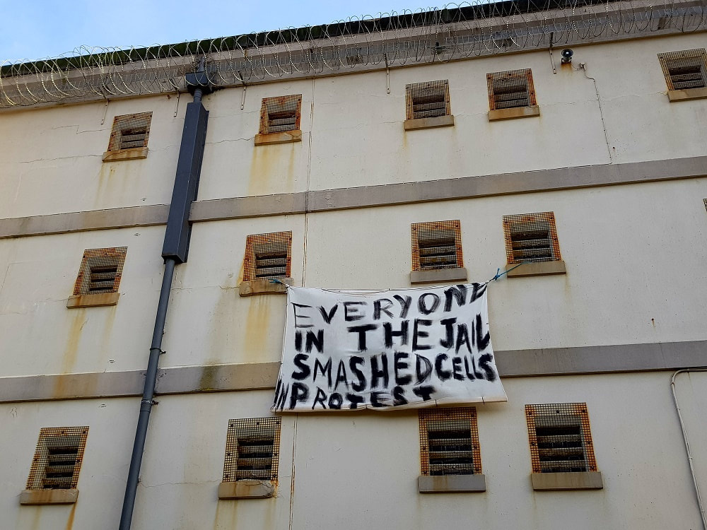 A beige building with a white banner hanging from one window reading 'Everyone in the jail smashed cells in protest'