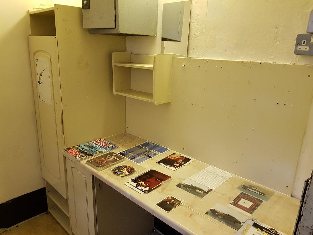 The interior of a prison cell with a cupboard, shelving and a desk with magazines on top