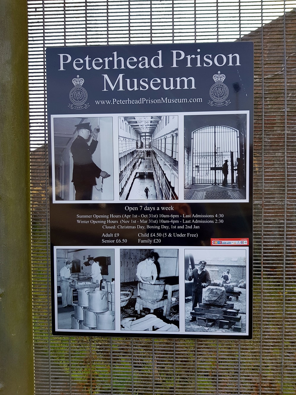 A black and white sign attached to a gate with details of Peterhead Prison Museum opening times and entry costs