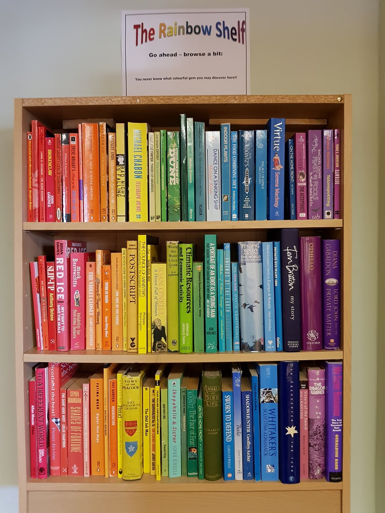 Three rows of shelves filled with multi-coloured books and a sign reading 'The Rainbow Shelf, Go ahead-browse a bit'