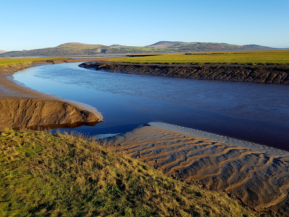 A river flowing through a silted estuary with green hills in the background
