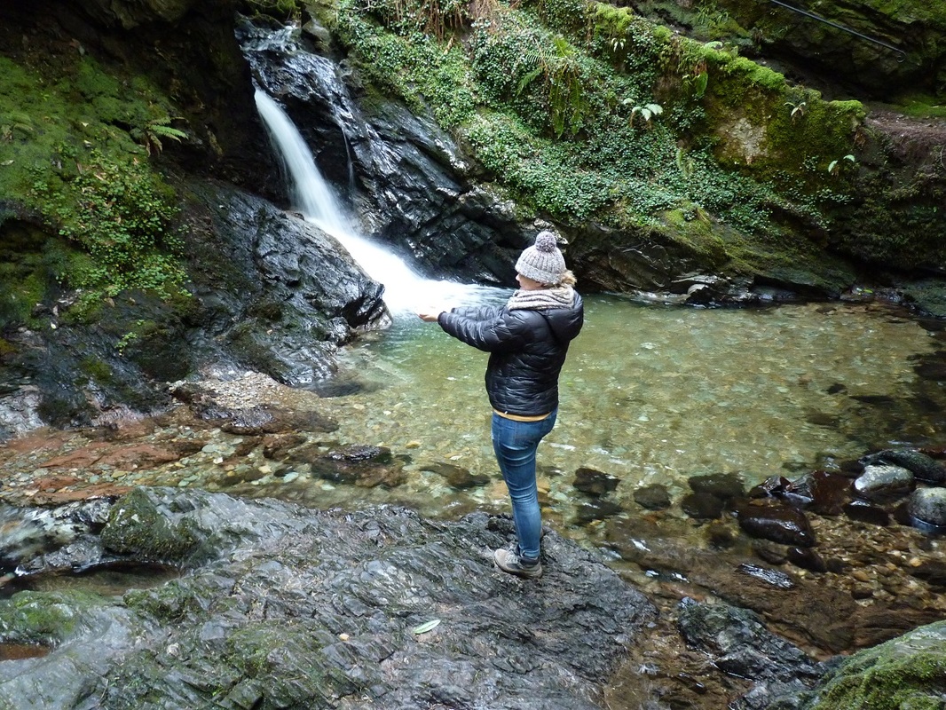 A woman standing next to a pool of water with her hands under a waterfall.