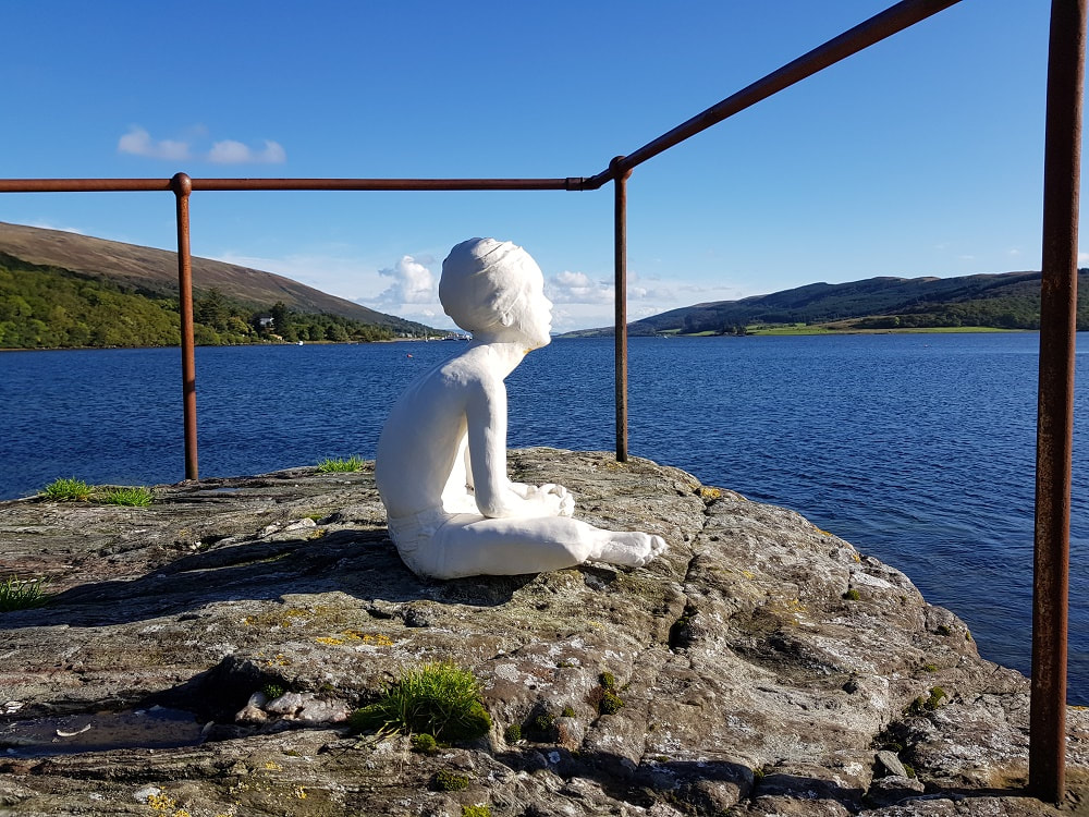 A white sculpture of a boy sitting on a rock looking at the water