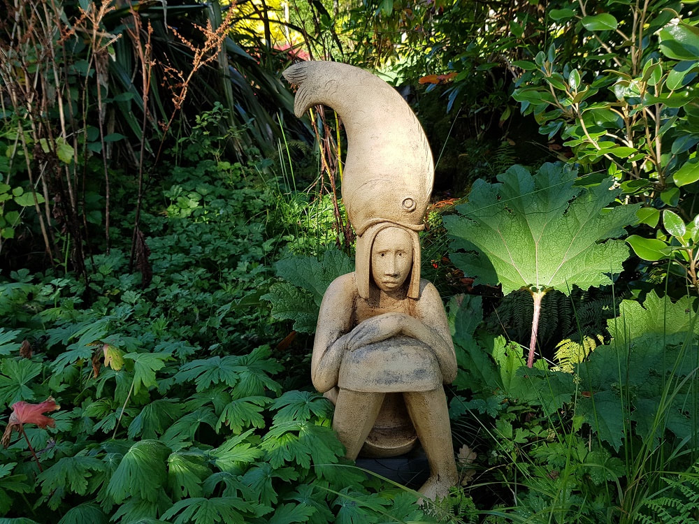 A wooden sculpture of a girl sitting in a garden with a fish on her head