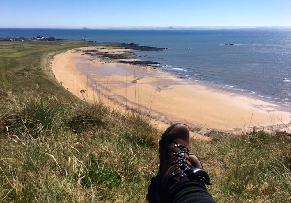 Someone sitting on a grassy hill overlooking a beach with only their walking boots showing