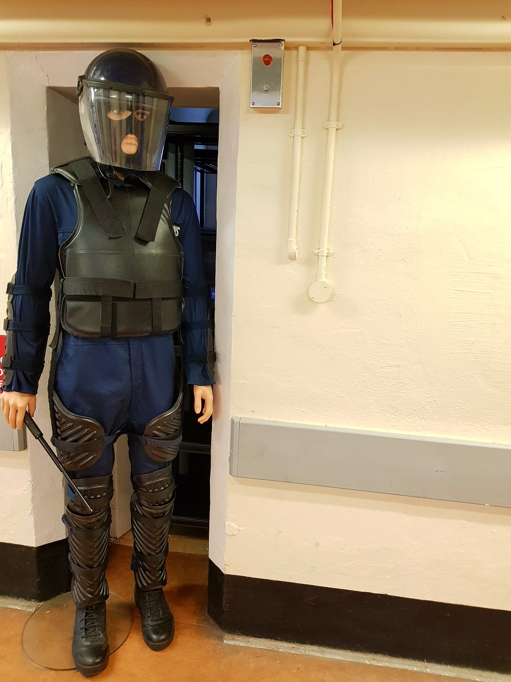 A mannequin carrying a truncheon, wearing blue overalls, a black helmet with visor, black body armour and padding on legs 
